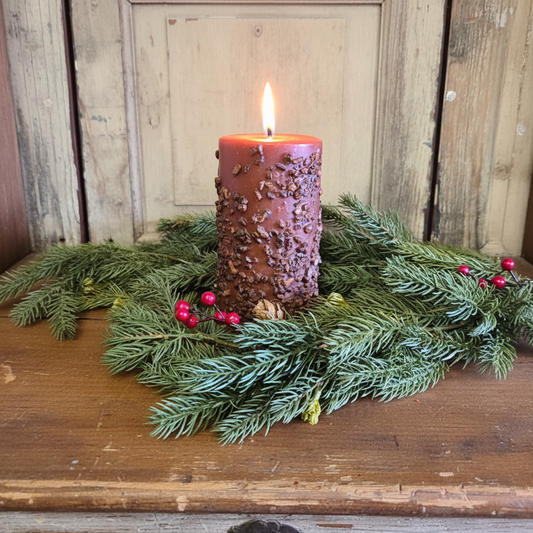 Decorative candle with textured surface and red berries on a bed of greenery against a wooden background