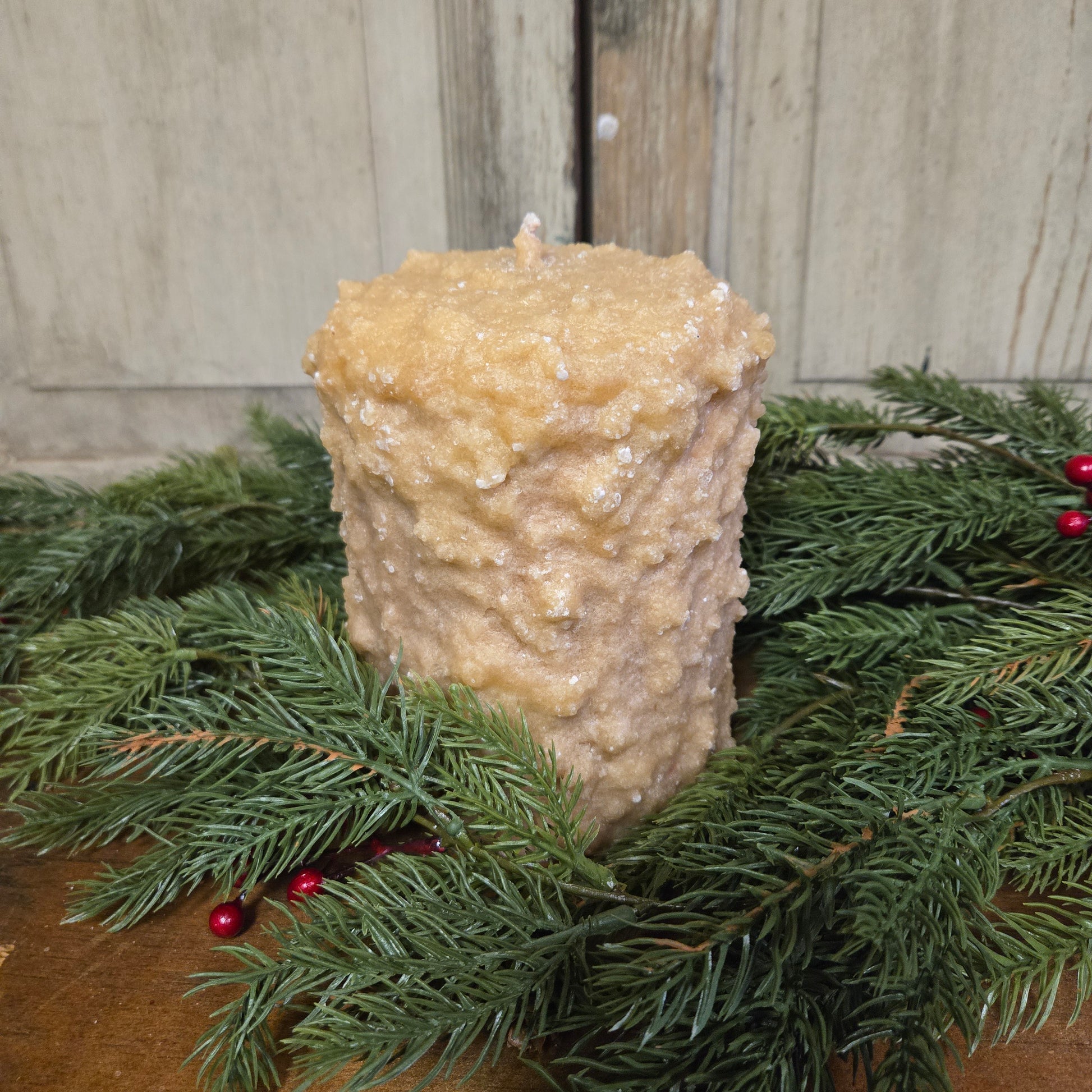 A large brown sugar candle with cranberry center, surrounded by green pine branches and red berries.