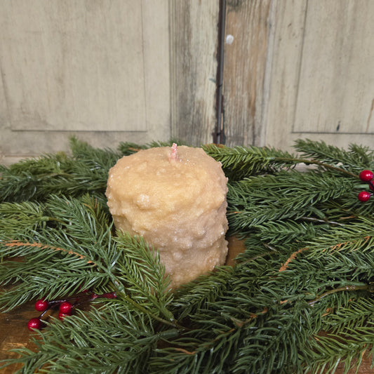 A cream colored candle with a bumpy texture surrounded by Christmas greenery