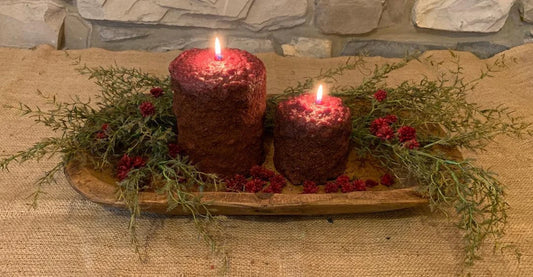 Two different size burning candles in a wood bowl with green sprigs