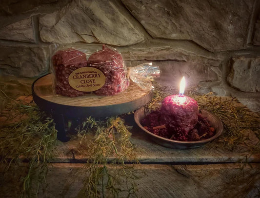 A two-pack of Cranberry Clove votive candles placed on a wooden platter, with one candle lit, surrounded by stones and a small branch on a wooden surface.