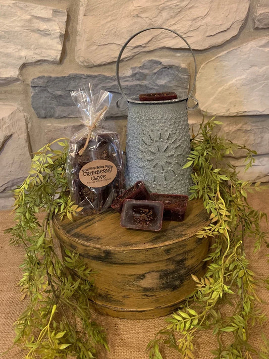 A set of cranberry clove wax melts displayed on a wooden platform with a decorative greenery backdrop. The wax melts are packaged in a clear bag and a metal container, and appear as dark brown or black candle pieces with a fragrant holiday scent.