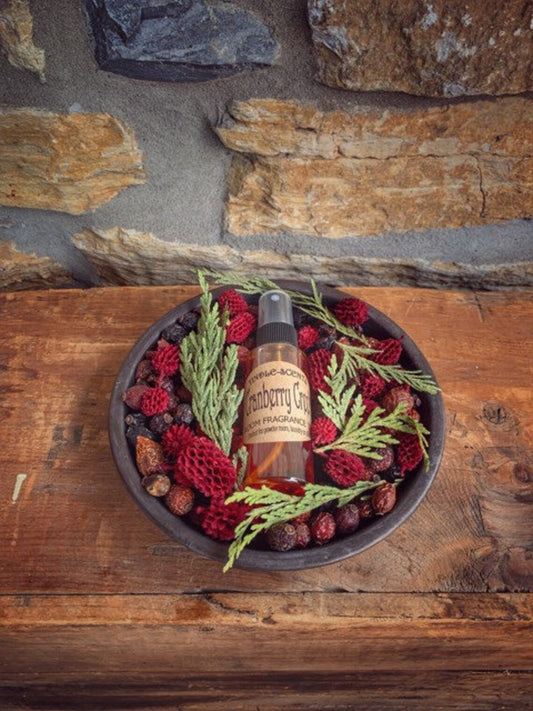 A metal pan with a bottle of oil and a sprig of greenery, surrounded by cranberries and potpourri on a wooden surface with a stone wall in the background.