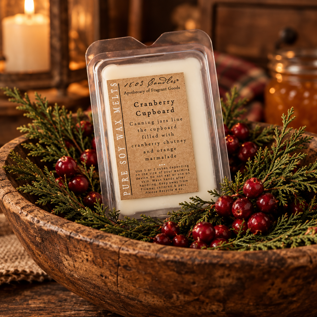 A packaged cranberry caboose scented soy wax melt from 1865 Apothecary of Fragrant Goods, displayed in a rustic wooden bowl filled with green pine branches and red berries.