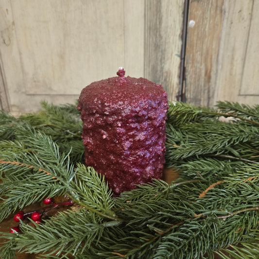 A small pillar candle with a cranberry-colored wax, placed on a pine branch, with a rustic wooden background.
