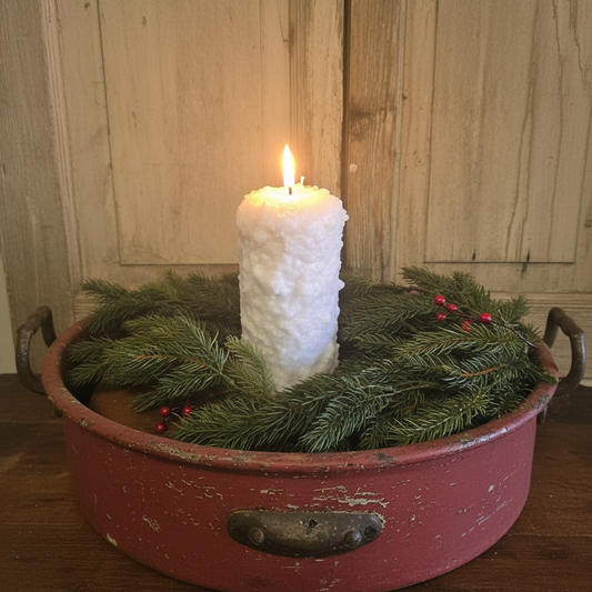 White textured candle in a red metal container with greenery against a wooden background