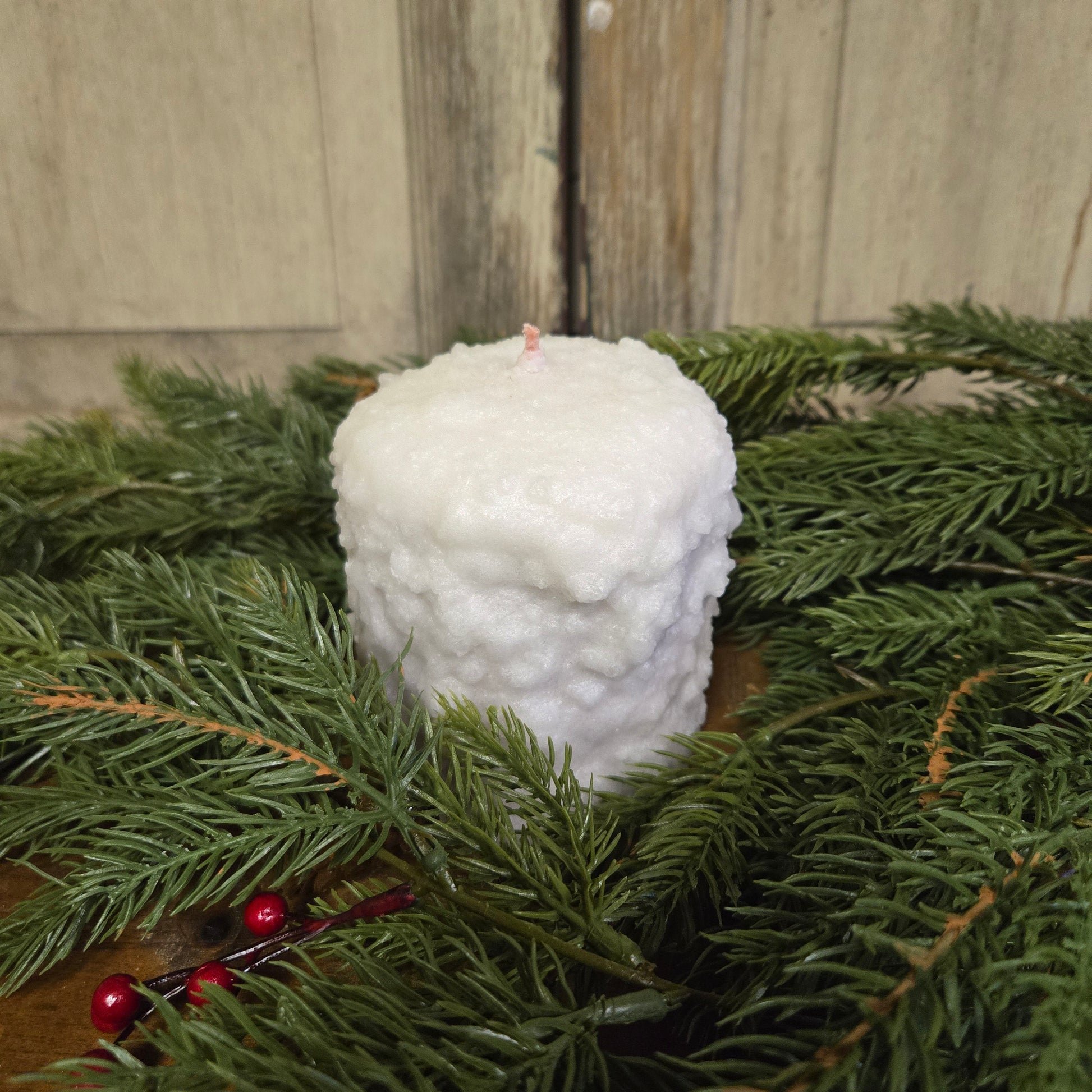 A small hearth candle with a cranberry center and sugar cookie frosting, surrounded by green pine branches and red berries on a wooden surface.