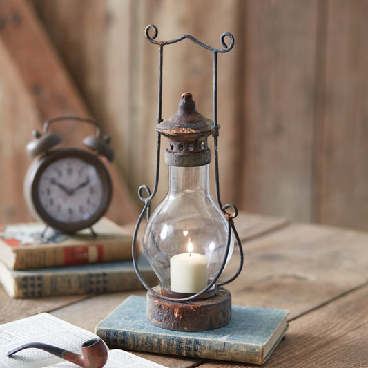 An antique-style metal and glass lantern with a lit candle inside, placed on a wooden surface next to an old clock and some books.