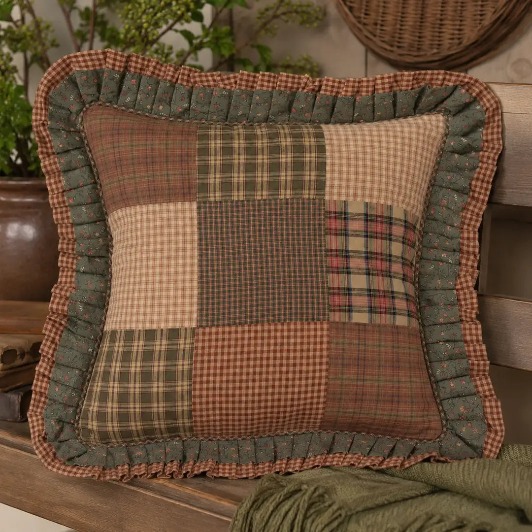 A square patchwork decorative throw pillow with a ruffled border, featuring various rustic plaid and checkered patterns in earthy tones of green, brown, beige, and red, styled on a wooden shelf with a green knit blanket and potted plants in the background.