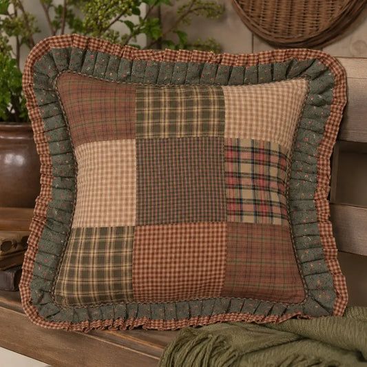 A square patchwork decorative throw pillow with a ruffled border, featuring various rustic plaid and checkered patterns in earthy tones of green, brown, beige, and red, styled on a wooden shelf with a green knit blanket and potted plants in the background.
