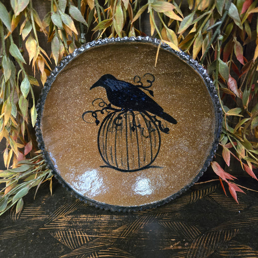 Round stoneware plate with a crow on a pumpkin vine on a textured wood surface surrounded by greenery