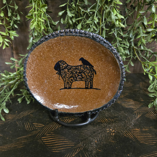 Brown ceramic dish with sheep and crow silhouette on a textured surface with greenery
