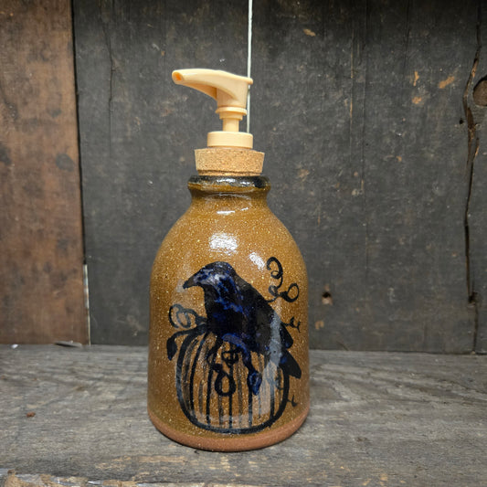 A handmade ceramic soap dispenser bottle with a brown speckled glaze, featuring a black painted raven perched on a birdcage design. The bottle has a cork stopper and a cream-colored plastic pump dispenser, set against a rustic dark wood background.