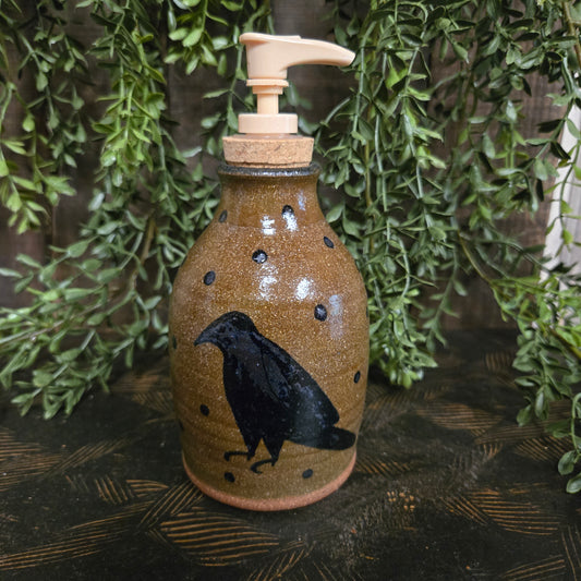 Ceramic bottle with pump and black bird design on a wooden surface with greenery.