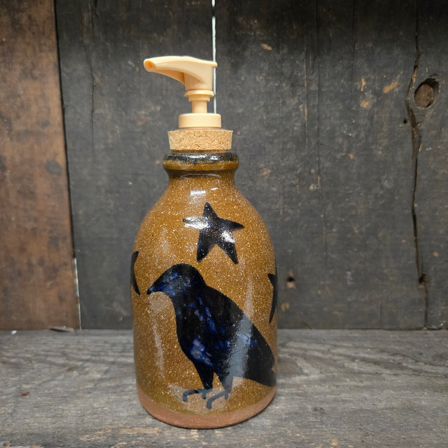 A handmade ceramic soap dispenser bottle with a brown speckled glaze, featuring a black raven and star design, a cork stopper, and a light wood pump, set against a rustic dark wood background.