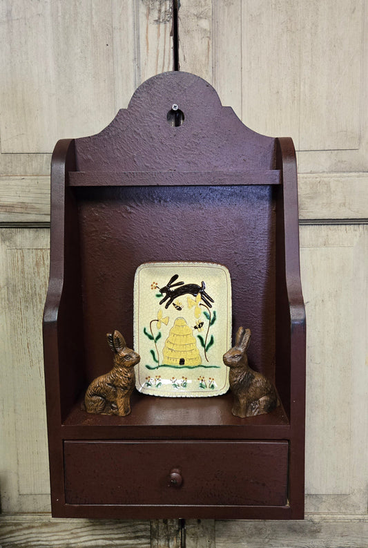 Small wooden shelf with a decorative panel and two small figurines against a wooden wall.