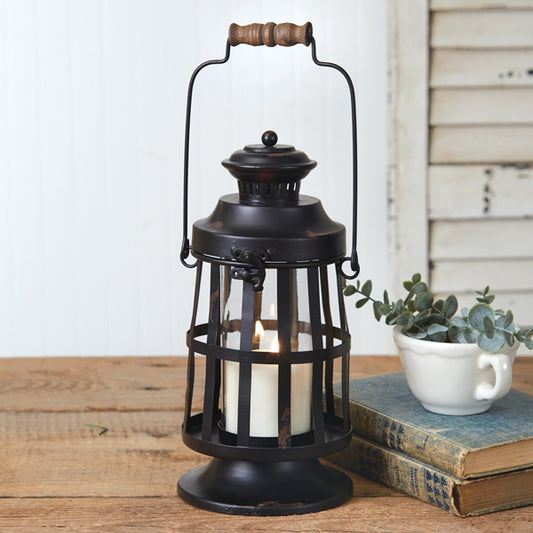 A black metal candle lantern with a glass chimney, featuring a flickering candle inside, placed on a wooden surface next to some books and a cup.