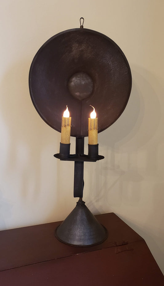 A double tin table light with a metal base and a round tin reflector, featuring two candles lit on top.