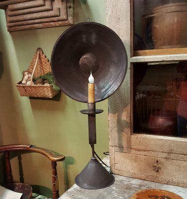 A tin table light with a cylindrical shape and a visible bulb, mounted on a stand, placed on a table.