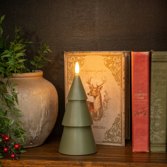 Green tree-shaped candle on a wooden surface with books and a vase in the background