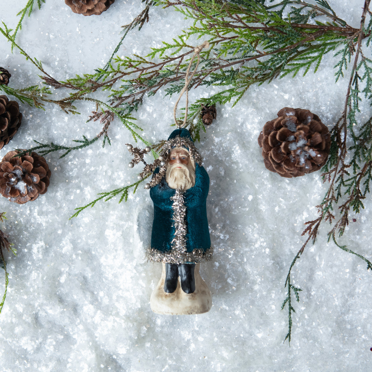 A deep ocean blue velvet Belsnickel ornament with a snowy backdrop, surrounded by pine cones and decorative greenery.