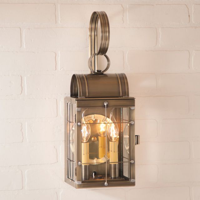 A double wall lantern in weathered brass with clear glass and a bronze-colored frame, mounted on a white brick wall, emitting light from within.