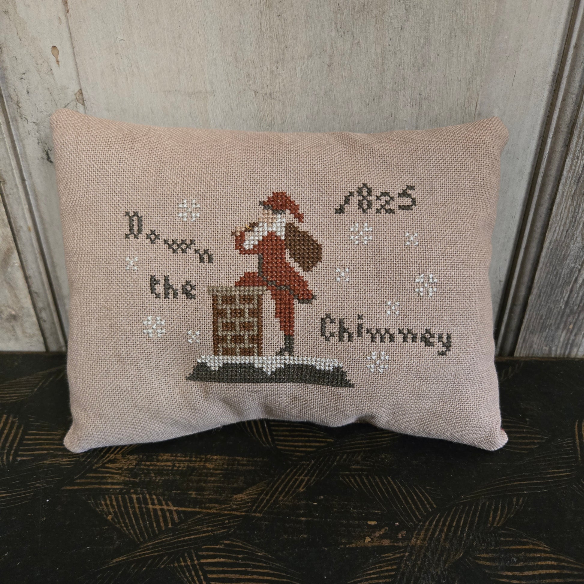 Decorative pillow with cross-stitched design of a santa and text, leaning against a textured wall.
