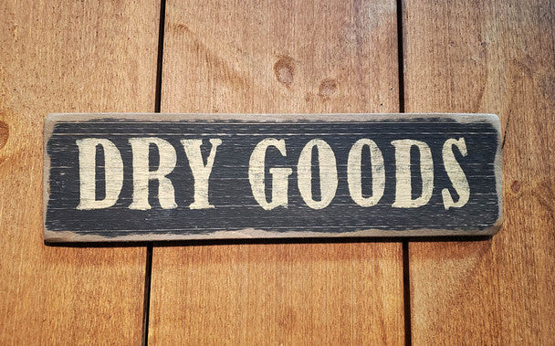 A wooden sign with the text 'Dry Goods' carved into it, placed on a wooden surface.