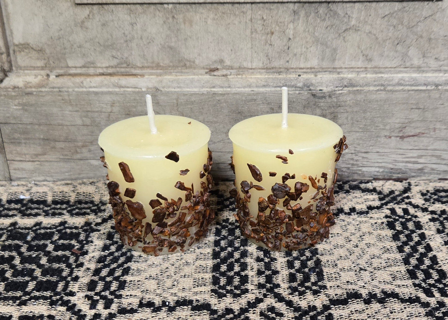 Two candles on a textured black mat with a knob handle, surrounded by a woven beige area rug. The candles are melted with visible wax drippings.