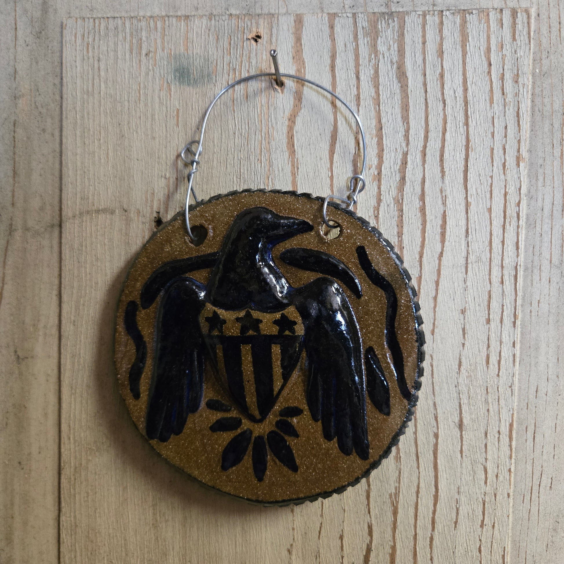 A round hanging ornament with a stoneware background and a glossy black eagle design with a shield motif, suspended by a silver wire hanger against a light wood plank background.