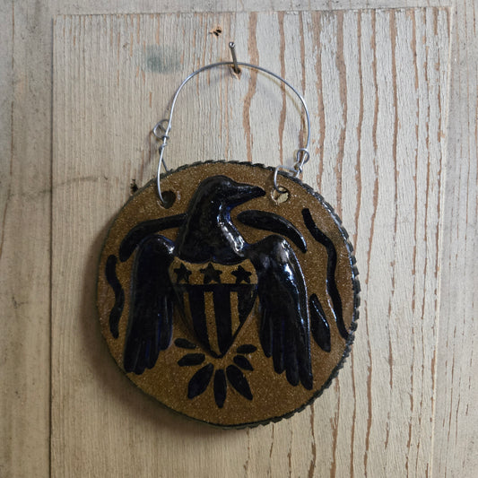 A round hanging ornament with a stoneware background and a glossy black eagle design with a shield motif, suspended by a silver wire hanger against a light wood plank background.