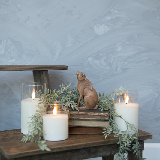 A brown resin bunny figurine placed on a wooden surface surrounded by three lit candles and decorative greenery.