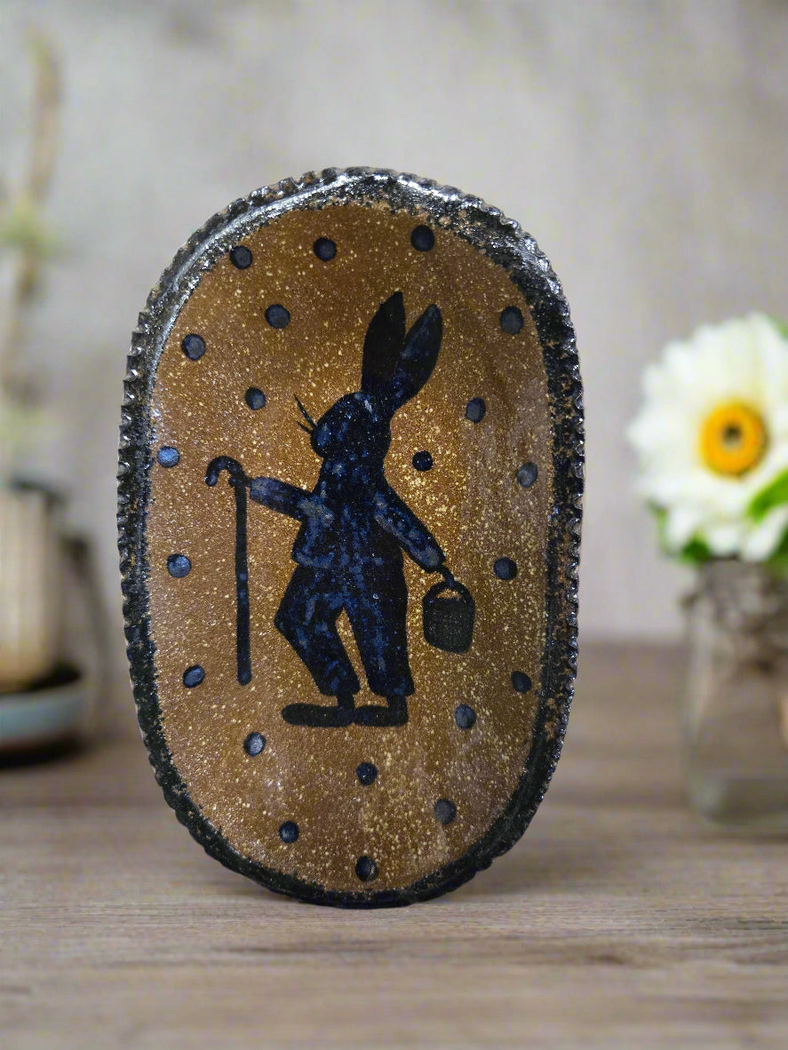 A medium oval stoneware plate with a brown glazed finish and a centered black and white decorative design, displayed on a wooden surface with a patterned fabric edge.