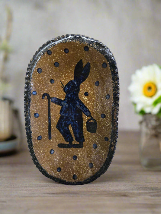 A medium oval stoneware plate with a brown glazed finish and a centered black and white decorative design, displayed on a wooden surface with a patterned fabric edge.