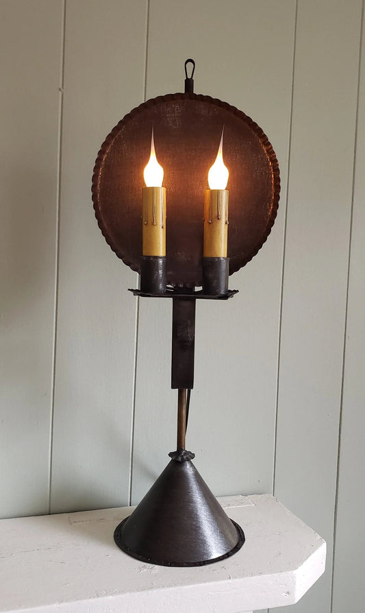 A rustic style table lamp with a round primitive finish shade and a cone base, featuring two candle-like lights.