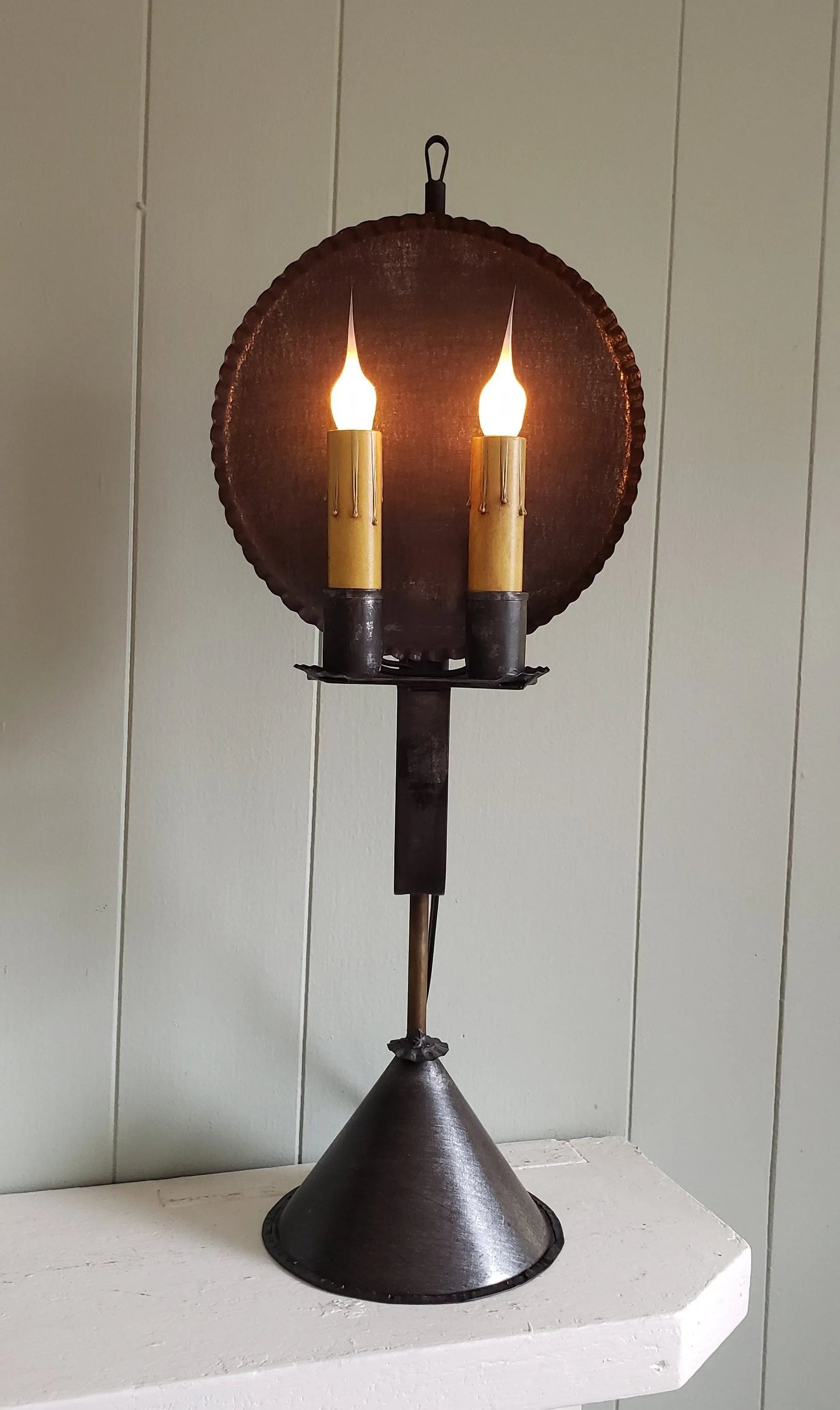 A rustic style table lamp with a round primitive finish shade and a cone base, featuring two candle-like lights.