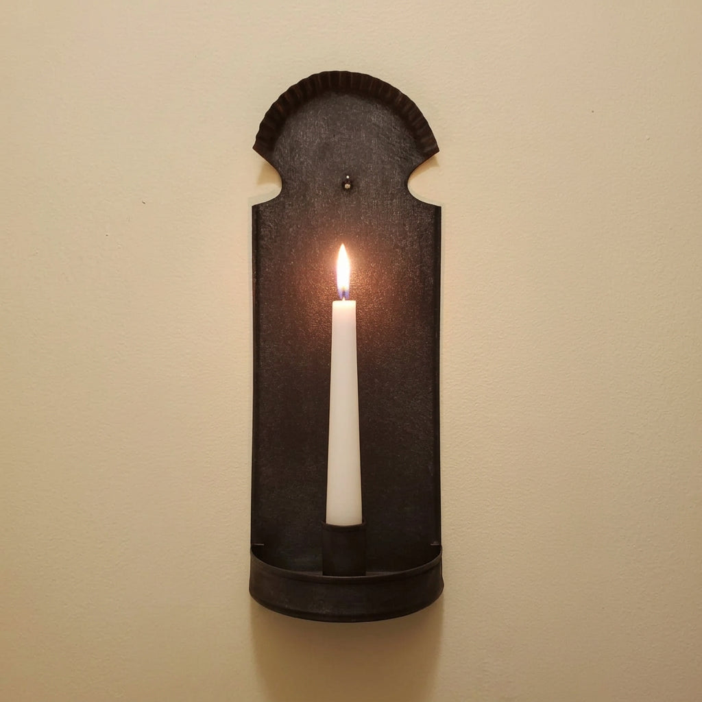 A dark metal wall-mounted candle holder with a scalloped top edge and a small cup at the bottom, shown against a plain beige wall.
