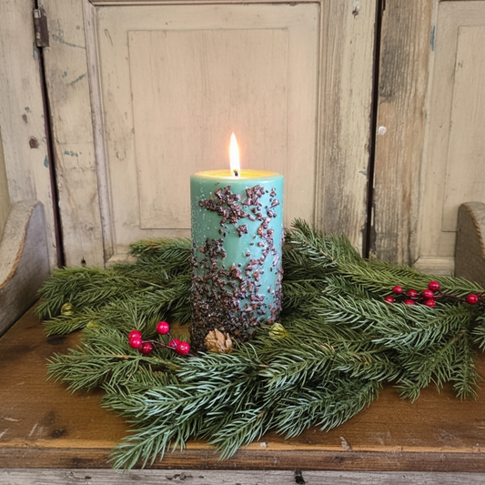 Teal candle with decorative elements on a bed of greenery against a wooden background