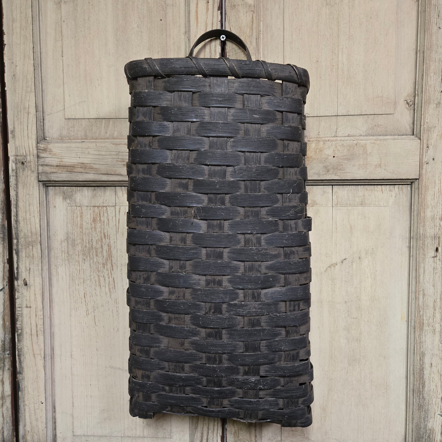 A tall black basket with a solid pattern, hanging on a wooden door