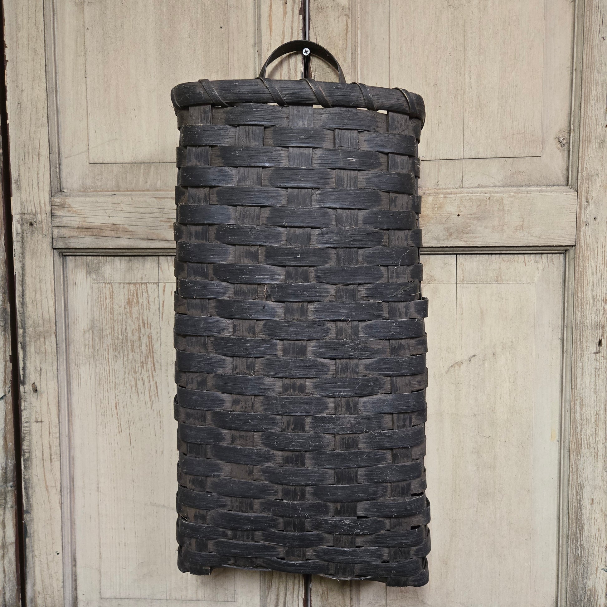 A tall black basket with a solid pattern, hanging on a wooden door