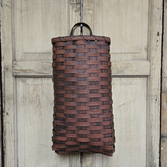 A tall, cylindrical door basket with a burgundy finish, hanging on a wooden door.