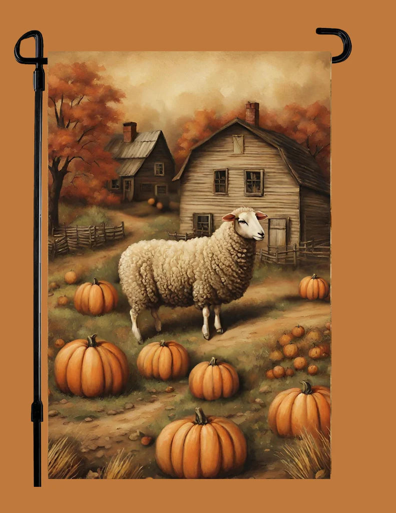 A vertical garden flag featuring an autumn farm scene with a sheep standing among pumpkins in front of rustic houses and trees, displayed on a black garden flag stand against a solid orange-brown background.