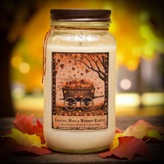 A Mason jar candle with a dark brown lid and a rustic autumn-themed label reading 'Leaves, Moss & Woody Earth.' The candle is photographed on a wooden surface with scattered red and yellow leaves, against a softly blurred warm outdoor background.