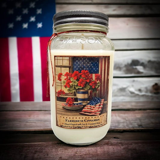 A mason jar candle with a 'Farmhouse Cinnamon' label, featuring a vintage-style lid, placed on a wooden surface with an American flag in the background.