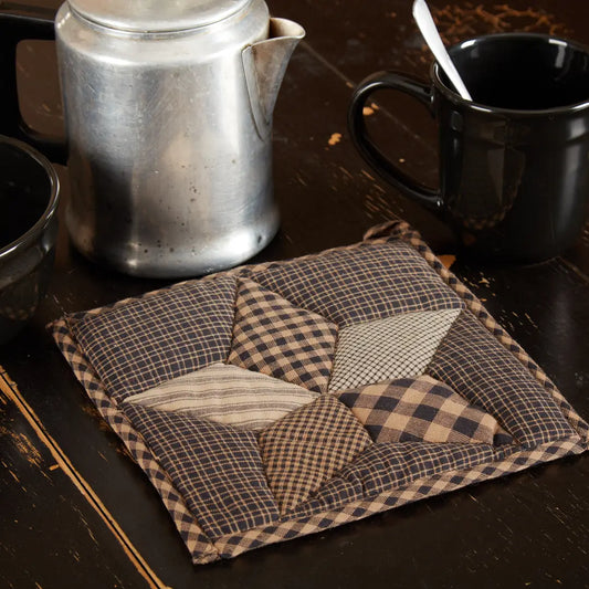 A square quilted fabric potholder with a patchwork star design in brown, beige, and black gingham patterns, displayed on a dark wooden table next to a metal coffee pot and a black mug.