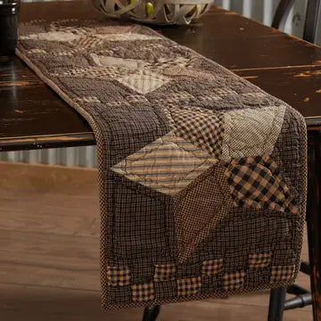 A rustic patchwork table runner with a star pattern in brown, beige, and black gingham fabrics, draped over a dark wooden dining table.