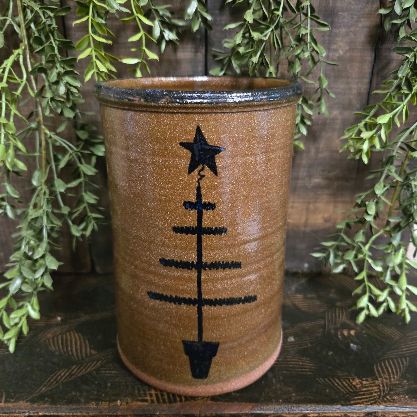 Stoneware crock with feather tree design on a wooden surface with green leaves.