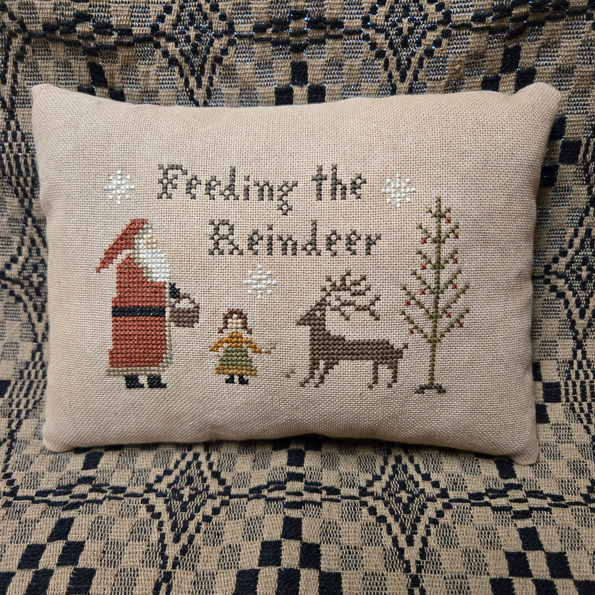 A square beige cross-stitch pillow featuring the text 'Feeling the Reindeer' and embroidered holiday figures including Santa, a reindeer, a girl, and a tree, displayed on a patterned couch.