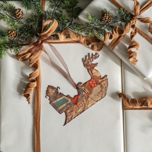 A vintage-style Christmas ornament shaped like a sleigh carrying a reindeer, decorated with distressed red, blue, and gold patterns, hanging on white wrapped gift boxes with brown ribbon bows and pine branches.