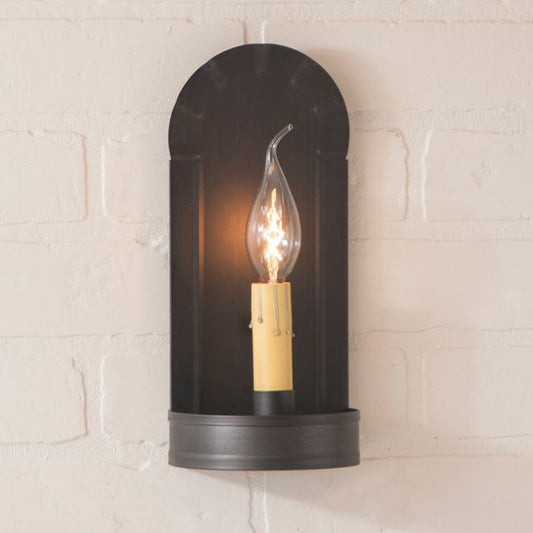 Black wall sconce with a lit bulb on a light beige brick wall.